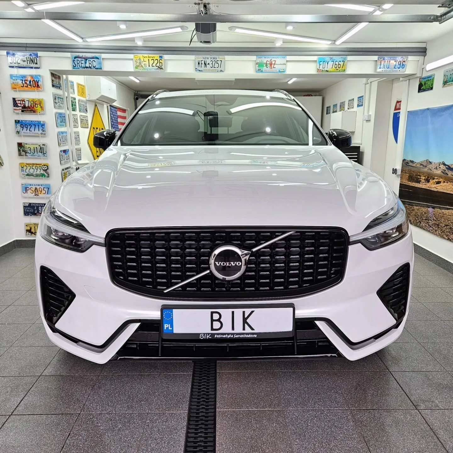 Przód białego Volvo XC60 na tle ściany z tablicami rejestracyjnymi z USA